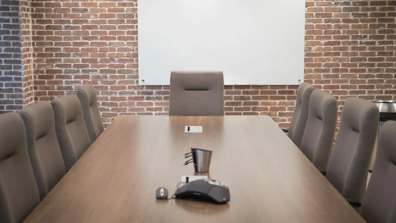 A long meeting table with chairs, a conference phone and a wall screen.