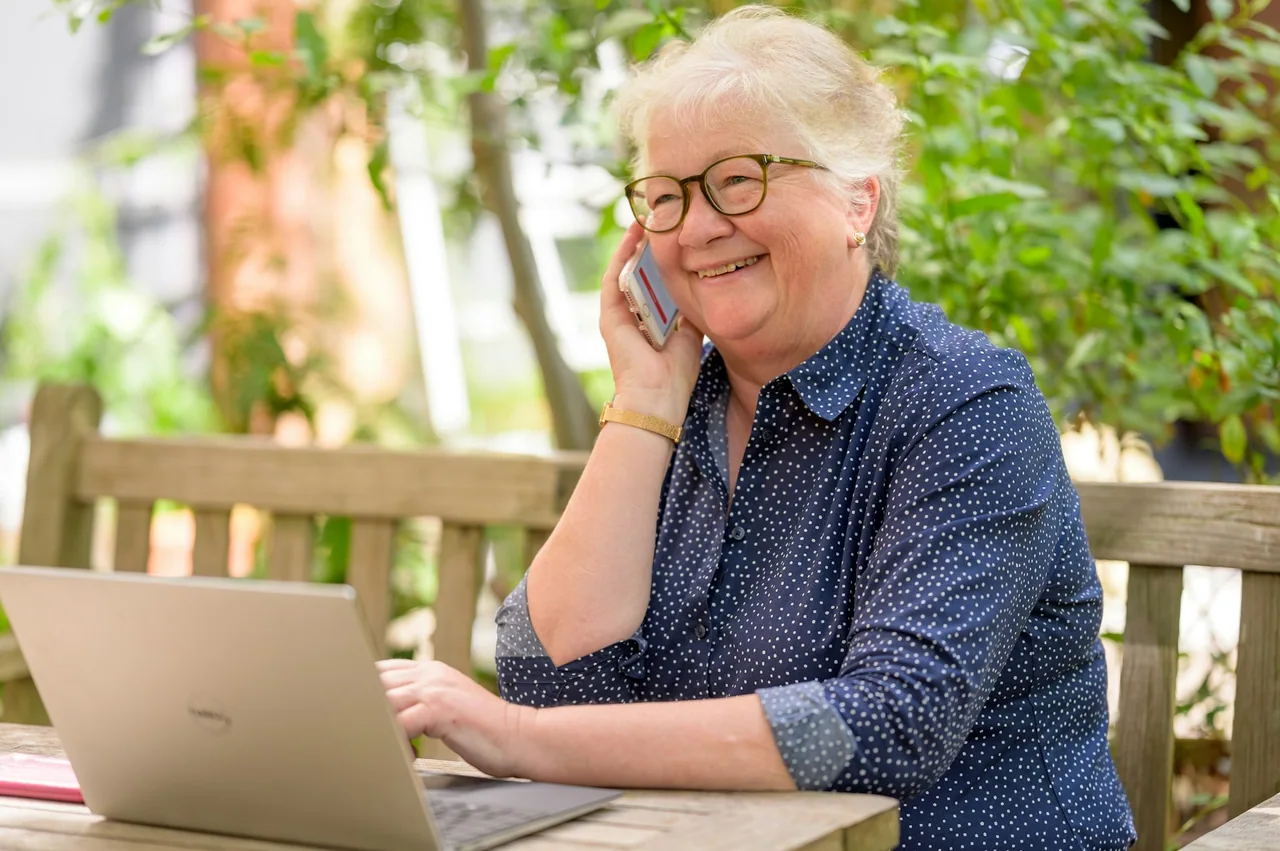 An older woman uses a laptop and speaks on a mobile phone