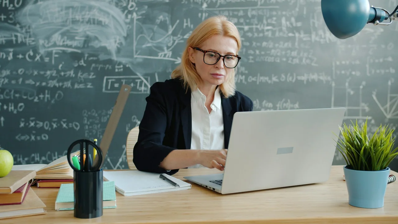 A teacher works on a laptop at a desk before a chalkboard